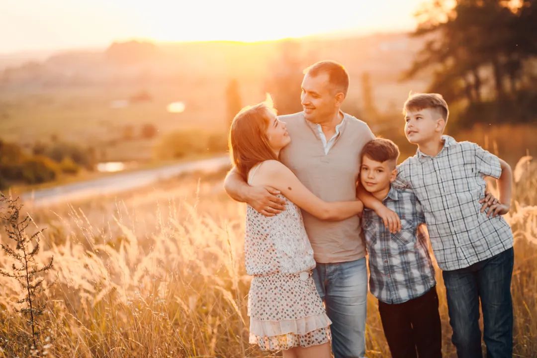 101057261_m A family of four stands together in a sunlit field, smiling and embracing, with golden grass and a picturesque landscape behind them.